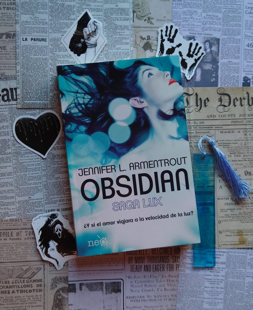 Imagen de la portada del libro Obsidian en el que se ve la cara de una joven de lado con colores azules rodeándola. También se ve un marcapáginas y varias pegatinas relacionadas con la historia.